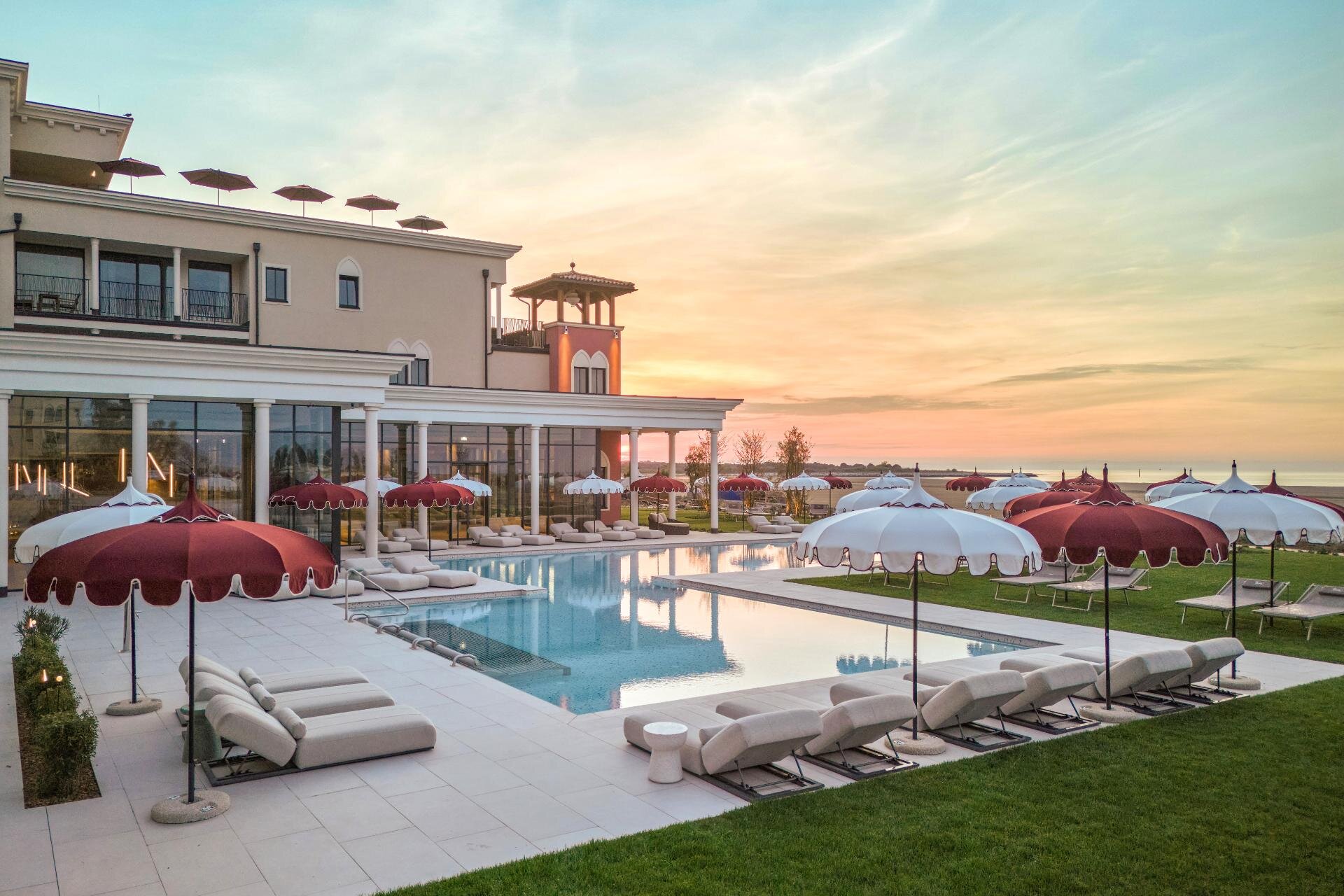 Outdoor swimming pool with red and white umbrellas in the resort garden in Caorle at sunset.
