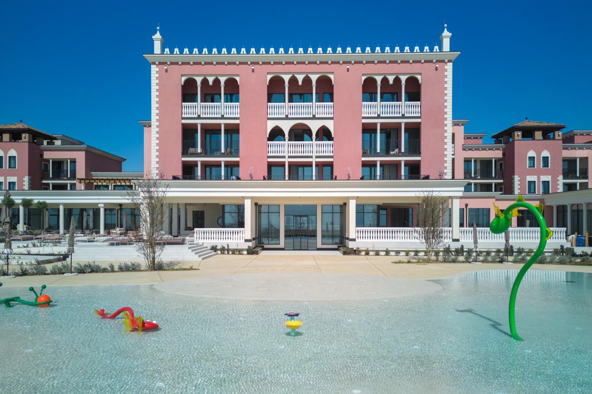 Children outdoor swimming pool area with water games in front of the pink building facade in Caorle.